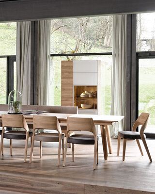 langer Holztisch mit Holzstühlen mit hellbrauner Ledersitzfläsche vor weißem Sideboard und Holzschrank