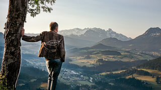 Mann lehnt sich mit den Arm an Baum und sieht auf Landschaftspanorama